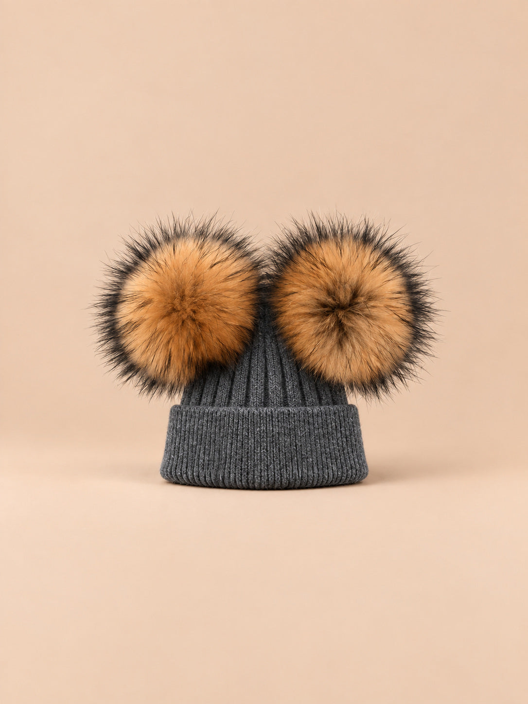 Bonnet double pompon, gris foncé, pompons naturels, vue de face sur fond pêche sable.